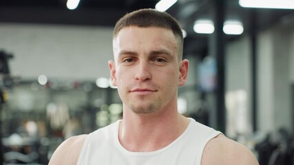 Portrait of gym goer standing beside fitness machine in fitness center, wearing tank top, looking down with calm smile while overhead lights reflect off equipment, showing focus and healthy lifestyle