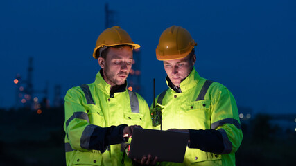 caucasian white engineers in safety uniforms and helmets using laptop and radio for site communication. Concept of teamwork, inspection, and operations at an industrial facility.Construction of Oil, 