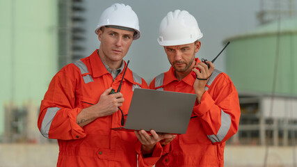 caucasian white engineers in safety uniforms and helmets using laptop and radio for site communication. Concept of teamwork, inspection, and operations at an industrial facility.Construction of Oil, 