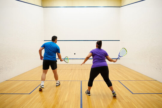 Fat man and woman playing squash on an indoor court, both holding rackets and facing the front wall.