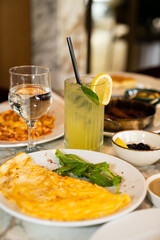 A glass of lemonade with ice, a lemon slice, fresh mint, and a straw is served with fried omelette, water and olives on a restaurant table.