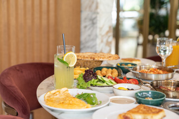 Close up view of a mixed breakfast table with various foods, including jams, cheese, fresh lemonade, butter, pastry, vegetables, olives, and an omelette, on a restaurant table.