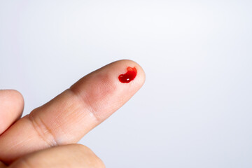 A light-skinned finger has a small puncture wound on its tip, from which a single, bright red drop of blood has emerged. The blood sits visibly against the plain white background.