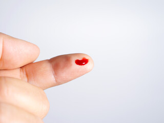 A light-skinned finger has a small puncture wound on its tip, from which a single, bright red drop of blood has emerged. The blood sits visibly against the plain white background.