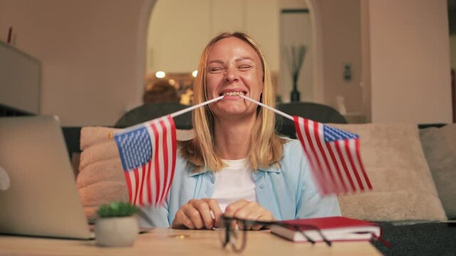 Energetic female gripping patriotic mini flags usa  between teeth, expressing national spirit during home workspace moment with laptop nearby