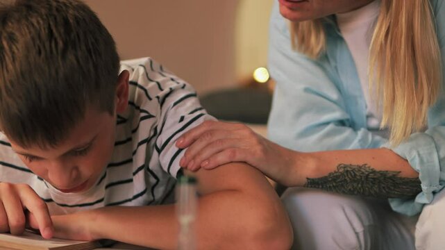 Caring mother supporting son's reading, resting hand affectionately on arm and fostering educational growth within home learning environment