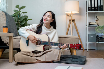 Asian girl sitting on floor playing the guitar at home, composing a song