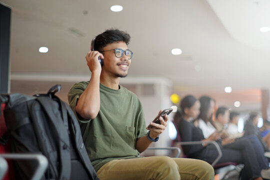 Smiling young Asian man using smartphone and headphones, chatting online, listening to music, while waiting in the waiting area and waiting to check in. - Powered by Adobe