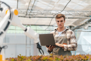 Male farmer using laptop in greenhouse to check the operation of robotic arm with AI system to control in farm care. Smart agriculture technology concept. Wireless robot for agriculture in smart farm. © Dee karen