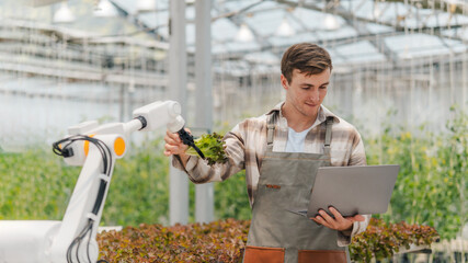 Male farmer using laptop in greenhouse to check the operation of robotic arm with AI system to control in farm care. Smart agriculture technology concept. Wireless robot for agriculture in smart farm.