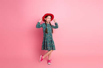 Adorable young girl in a printed dress posing with charm wearing a red hat in front of a pink background
