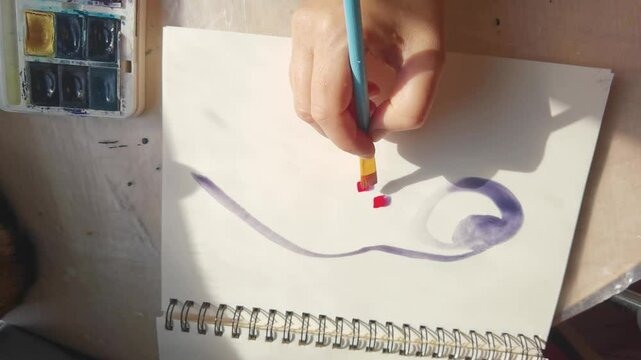 Artist's hand painting abstract design with watercolors in sunlit studio.