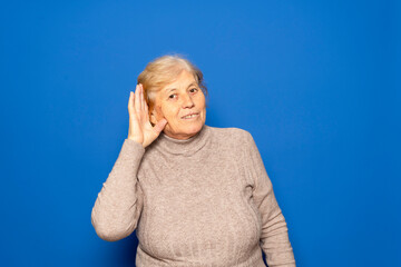 Caucasian woman in her 70s wearing a dark beige sweater putting her hand to her ear because she can't hear the most interesting gossip, isolated on blue background