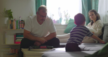 Obraz premium Father playing toy train with young boy at coffee table, mother smiling on sofa with small white dog, cheerful family bonding in cozy indoor living room environment