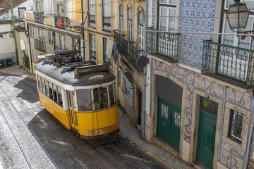 Historic yellow tram navigating a narrow, cobbled street in Lisbon, Portugal, with colorful tiled...