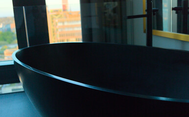 Black oval modern bathtub in a hotel room with a window and a view of a city in the background.