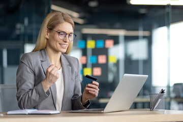 A smiling businesswoman in a blazer celebrates with a credit card while using her laptop, possibly online shopping.