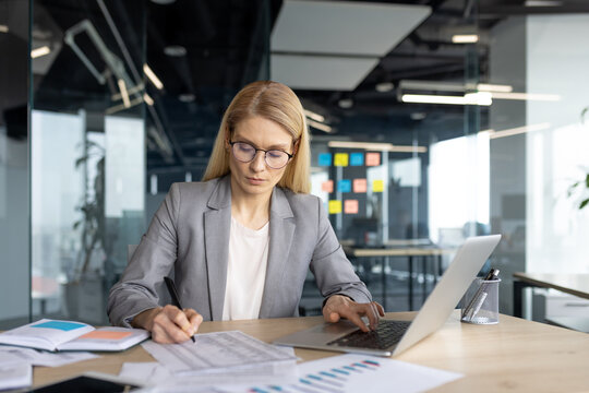 A businesswoman in a modern office setting is focused while working on a laptop and reviewing documents at her desk, analyzing data.