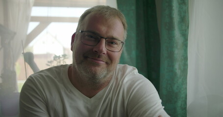 Man smiling warmly indoors, seated near a window with arms crossed, exuding friendliness and calm...