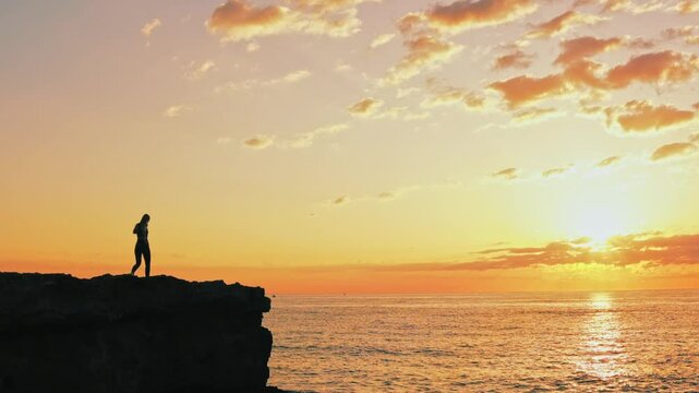 Muscular silhouetted female leaping from rocky cliff edge, diving toward golden horizon, expressing boundless freedom and adventurous spirit
