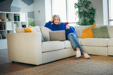 Obraz premium Mature woman relaxing on a cozy sofa at home with a laptop while enjoying a glass of red wine in a bright interior