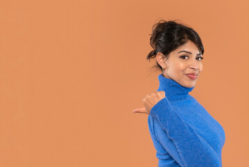 Woman in a blue turtleneck sweater gestures with her thumb over her shoulder against brown...