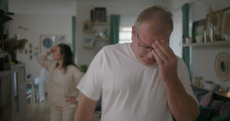 Obraz premium Man covering face with hands in frustration during argument, woman in background in living room, dramatic relationship conflict, focus on emotional struggle