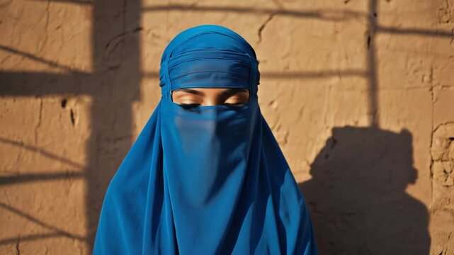 A woman draped in a stunning blue niqab, standing before a textured wall with sunlight casting intriguing shadows, creating a captivating scene of cultural significance and serene contemplation