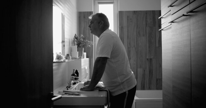 Man leaning on sink in black and white, deep in thought, reflective expression conveying life crisis and crossroads, serene bathroom setting with wooden details