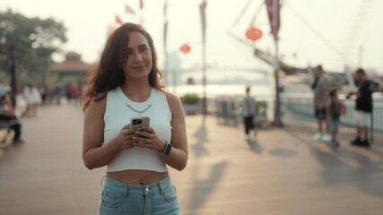 Portrait of happy young Latin woman holds smartphone walking along embankment. Pretty Hispanic female sends message on cellphone strolling past modern yachts in touristic port