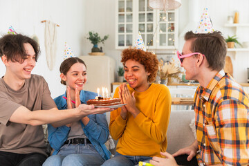 Make a wish. Woman wearing party cap blowing out burning candles on birthday cake. Happy Birthday party. Group of friends wishes girl happy birthday. People celebrating birthday with party at home
