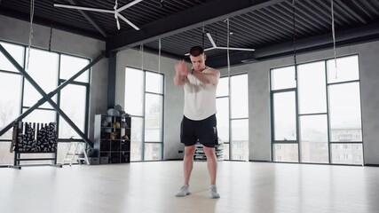 Body built man performing stretching exercise in spacious gym room with large windows and high ceiling, wearing white tank top and black shorts, preparing muscles for intense workout