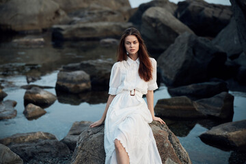 Serene woman in white dress sitting on oceanfront rock gazing at the sea with rocky background