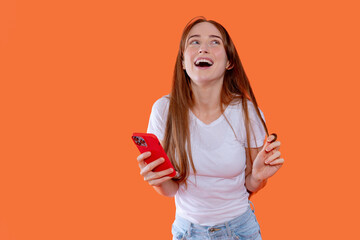 Young woman joyful with smartphone in hand against bright orange background while expressing excitement