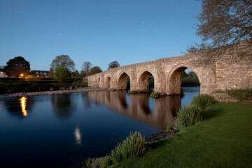 Fototapeta premium Stone bridge arches serenely reflect on moonlit waters, evoking Beltane whispers and hidden Celtic wanderlust