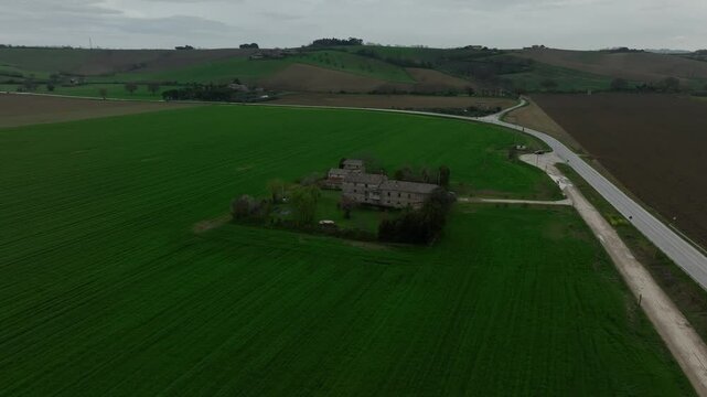 Drone flies over a lush green field in Italy, slowly approaching a charming house standing alone in the center of the landscape.