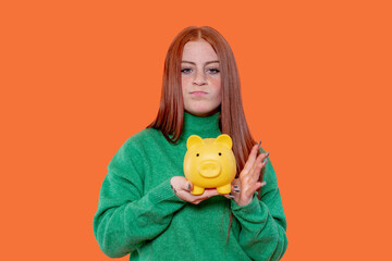 Young woman holding a yellow piggy bank with a displeased expression against a bright orange...