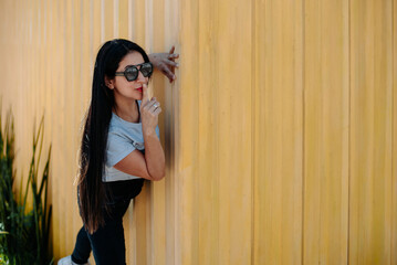 Obraz premium Confident woman with sunglasses poses near a yellow wall, making a silence gesture with her finger on her lips. Stylish, expressive and ideal for lifestyle or fashion themes.