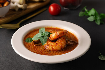 Fresh aromatic hot Thai soup Tom Yum with mushrooms, shrimps and different spices on black stone background close up. Tasty food concept.