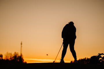 Silhouette of a female golfer hitting a ball at sunset, with club extended and golf bag nearby, against a vibrant orange sky on a peaceful course..