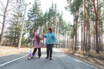 Fototapeta premium Little girl riding scooter while walking with her grandmother in the forest on a sunny day
