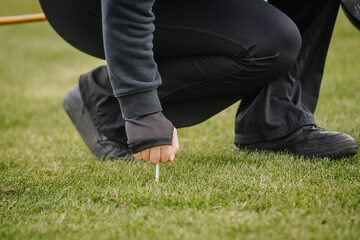 Close-up of a golfer kneeling to place a golf ball on a white tee on green grass, wearing black...