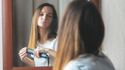 Young woman using a hair iron and doing a hairstyle looking at the mirror in the morning at bedroom...