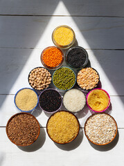 Assorted Grains and Legumes in Bowls Arranged in Pyramid Shape on White Wooden Background. Colorful grains, legumes, and pasta arranged in a pyramid shape on white wood under natural light.