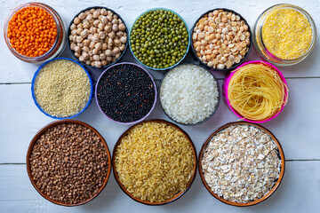 Top View of Various Grains, Legumes, and Pasta in Bowls on White Wooden Table. Assorted grains, legumes, and pasta displayed in colorful bowls on a white wooden surface, top view.