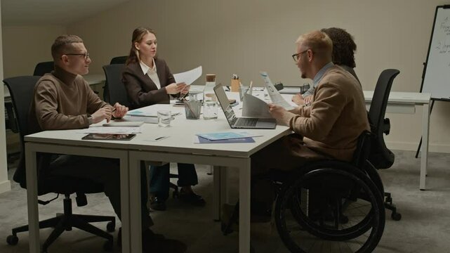 Push in camera movement on male team lead in wheelchair handing out documents with current agenda for discussion to colleagues gathered at desk in boardroom