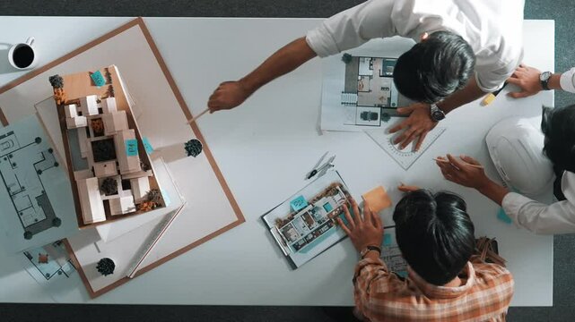 Group of skilled designer looking at model while discussing about building. Top view of architect engineer explain idea while talking and writing blue print scattering at meeting table. Alimentation.