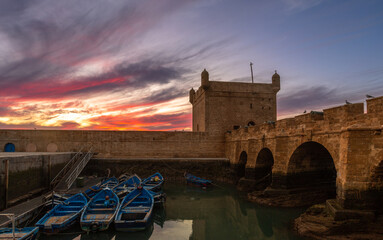 Blue fishing boats in a harbor with foretress bridge and Port of Essaouira Sqala, Portuguese castle in a sunset lights, Essaouira, Morocco