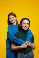 Fototapeta premium Portrait of two cheerful sisters hugging and smile looking at camera, isolated over yellow background