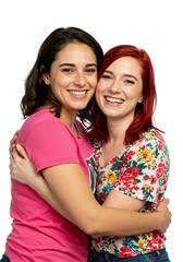 Two cheerful young adult women, one with dark hair in a pink shirt and one with red hair in a floral top, embrace warmly and smile happily at the camera against a transparent background.